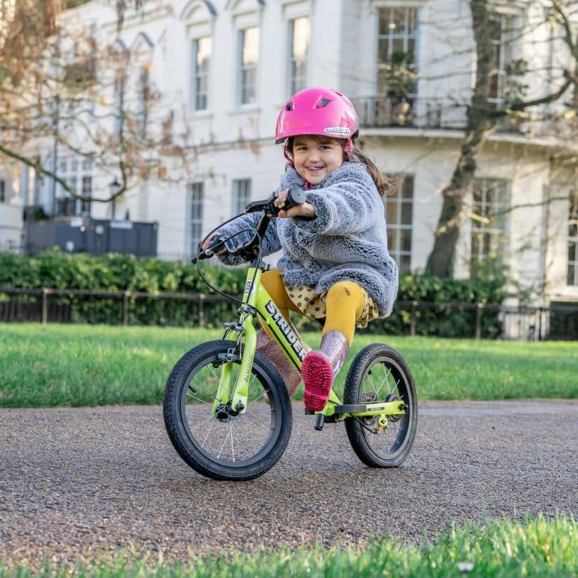 A girl in a pink bicycle helmet rides a green Strider 14x Sport bike in balance mode through a city park